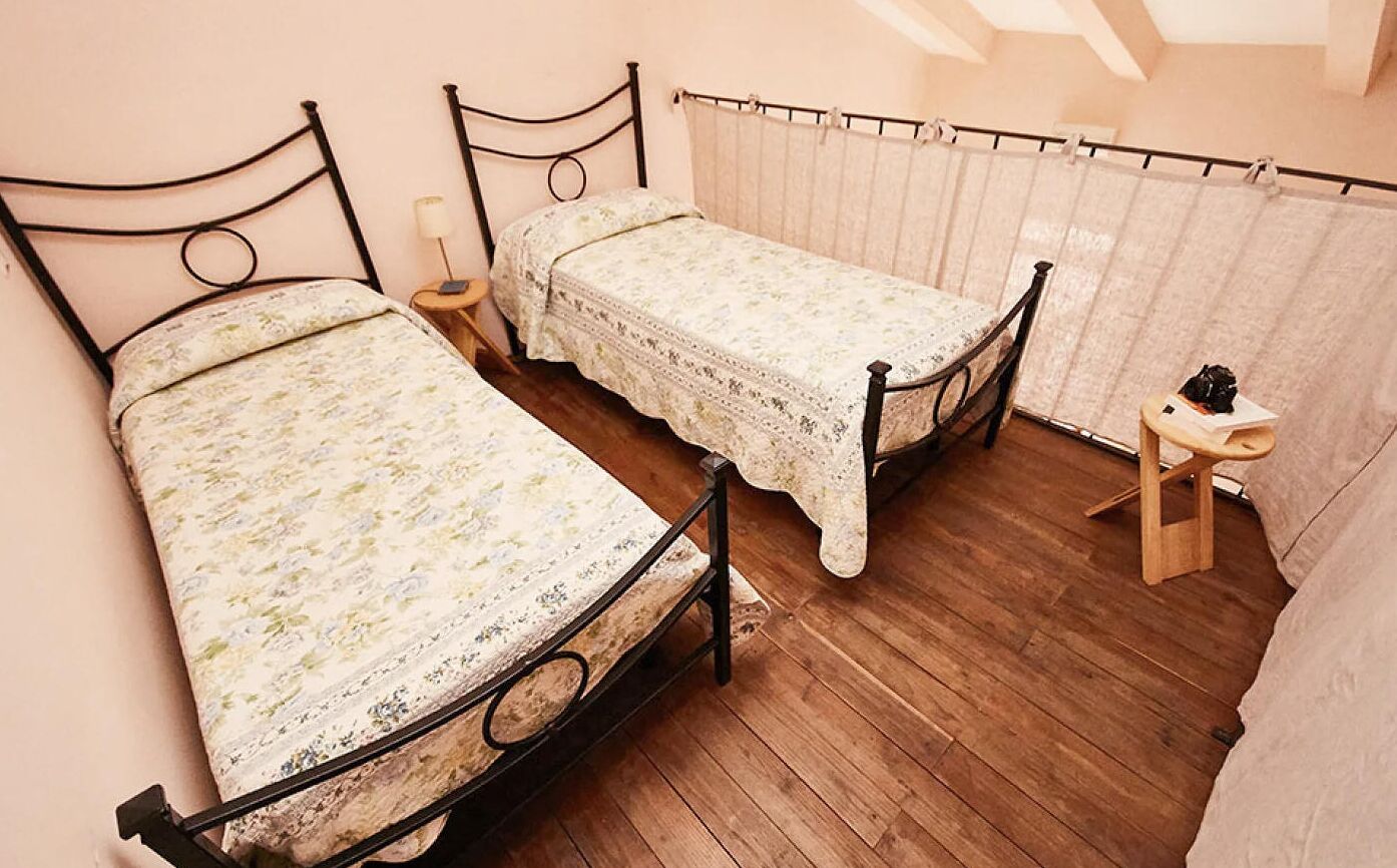 Een slaapkamer met twee eenpersoonsbedden, een houten vloer, een klein nachtkastje met een lamp en een raam met gordijnen.