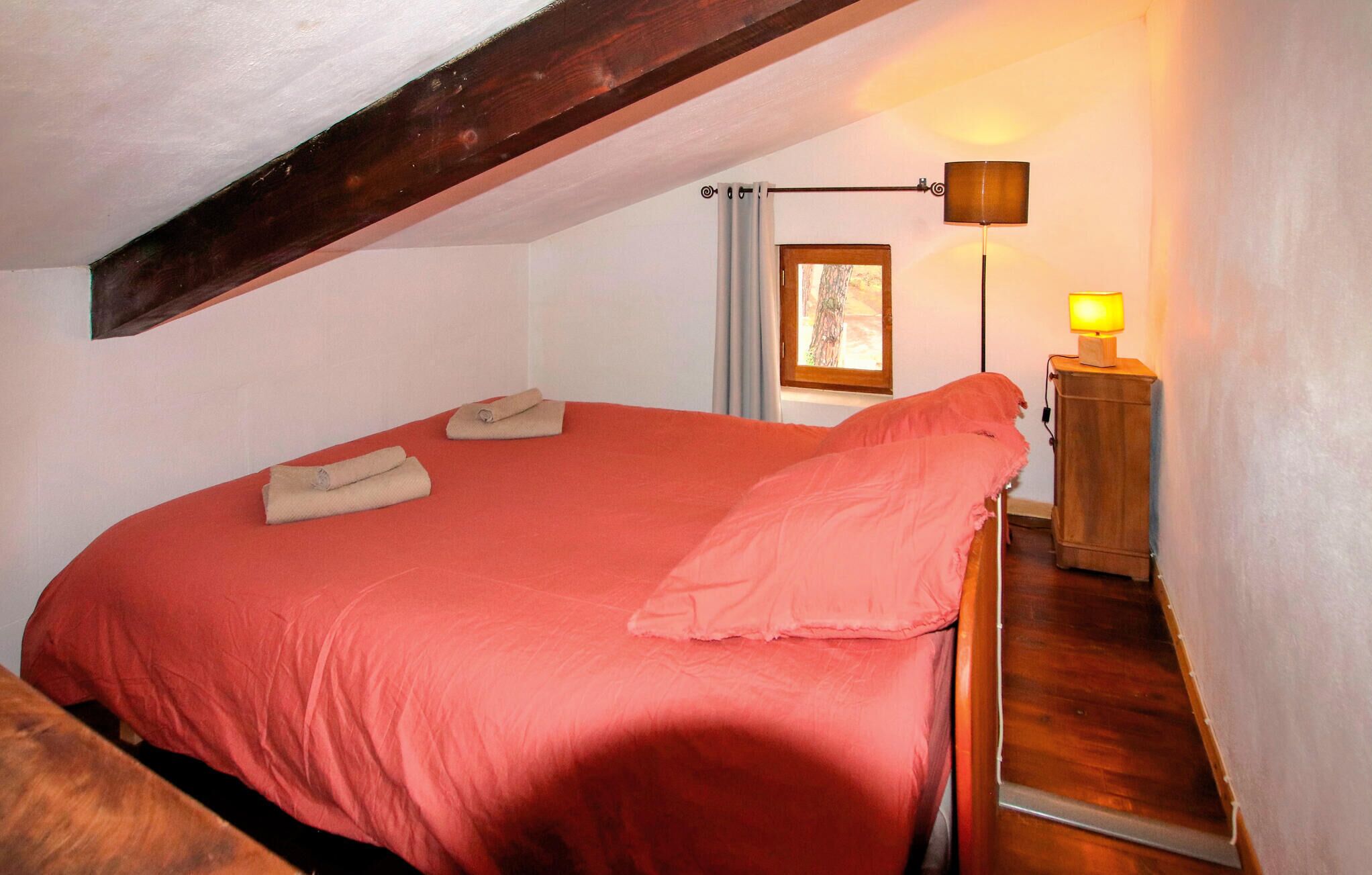 A bedroom with a red bedspread, a wooden headboard, a nightstand with a lamp, and a window with curtains.