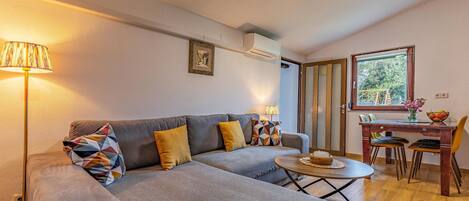 A cozy living room with a grey sofa, two lamps, a small table with a candle, and a window with a view of trees.