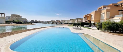 A swimming pool with a clear blue water, surrounded by a paved area and modern buildings in the background.