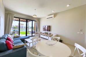A modern living room with a sofa, dining table, and chairs, large windows with curtains, a flat-screen TV, and an air conditioning unit. - Xuru Stays I Homey 3 BDR Townhouse @ Tawila El Gouna (El Gouna)