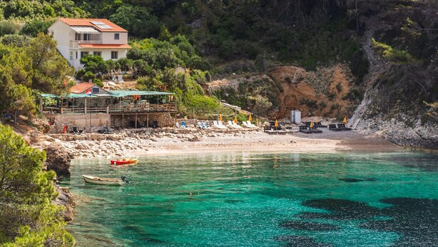 Un hôtel situé sur la côte, doté d’une terrasse donnant sur une plage, avec un bateau amarré près du rivage et une falaise rocheuse en arrière-plan.