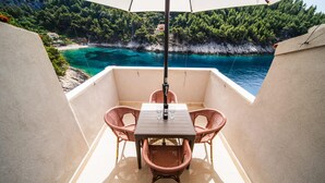 Un balcon agrémenté de fauteuils en osier et d’une table, offrant une vue sur une mer turquoise et une côte bordée de forêts.