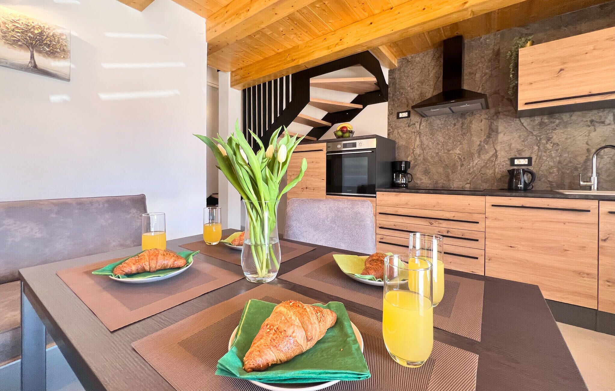 Een moderne keuken met houten kastjes, een inbouwoven en een spatwand van natuursteen. Er staat een eettafel met twee borden croissants en glazen jus d'orange.
