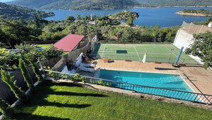 En pool med en tennisbana intill, omgiven av grönska och med berg i bakgrunden.