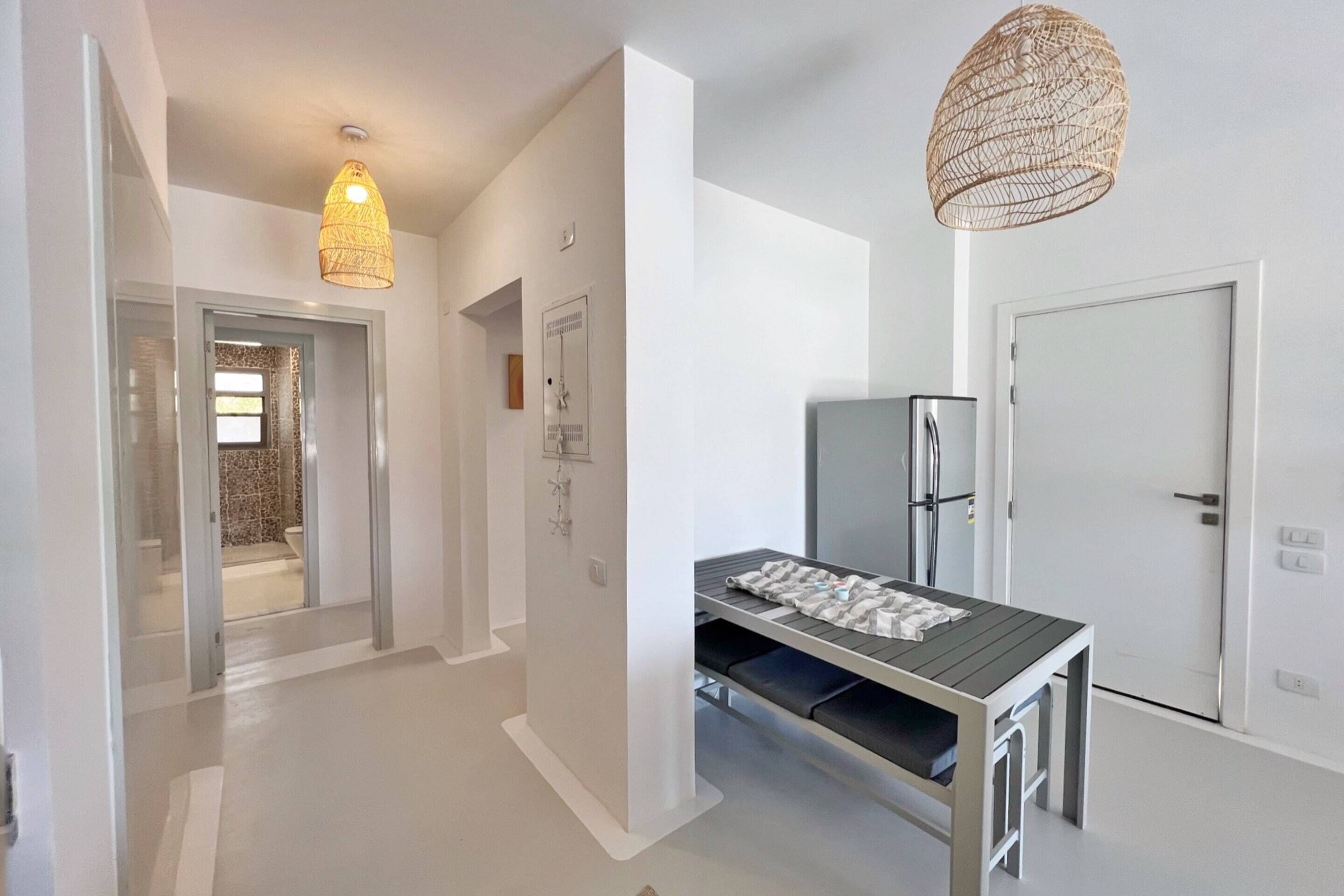 A modern, minimalist hallway with a table and bench, a refrigerator, and a hanging light fixture.