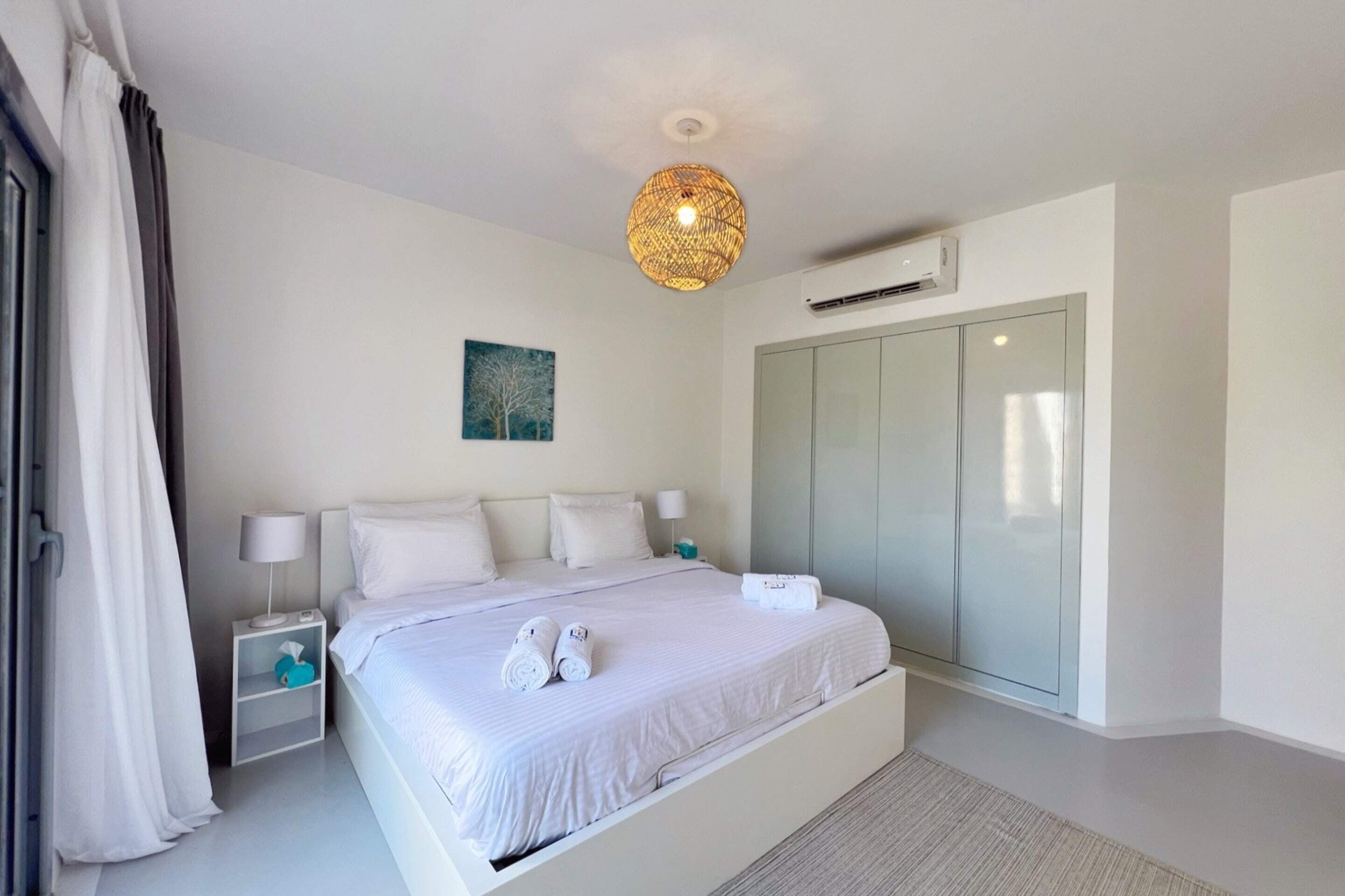 A modern bedroom with a large bed, bedside tables, a hanging light fixture, an air conditioning unit, and a wardrobe.