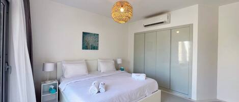 A modern bedroom with a large bed, bedside tables, a hanging light fixture, an air conditioning unit, and a wardrobe.