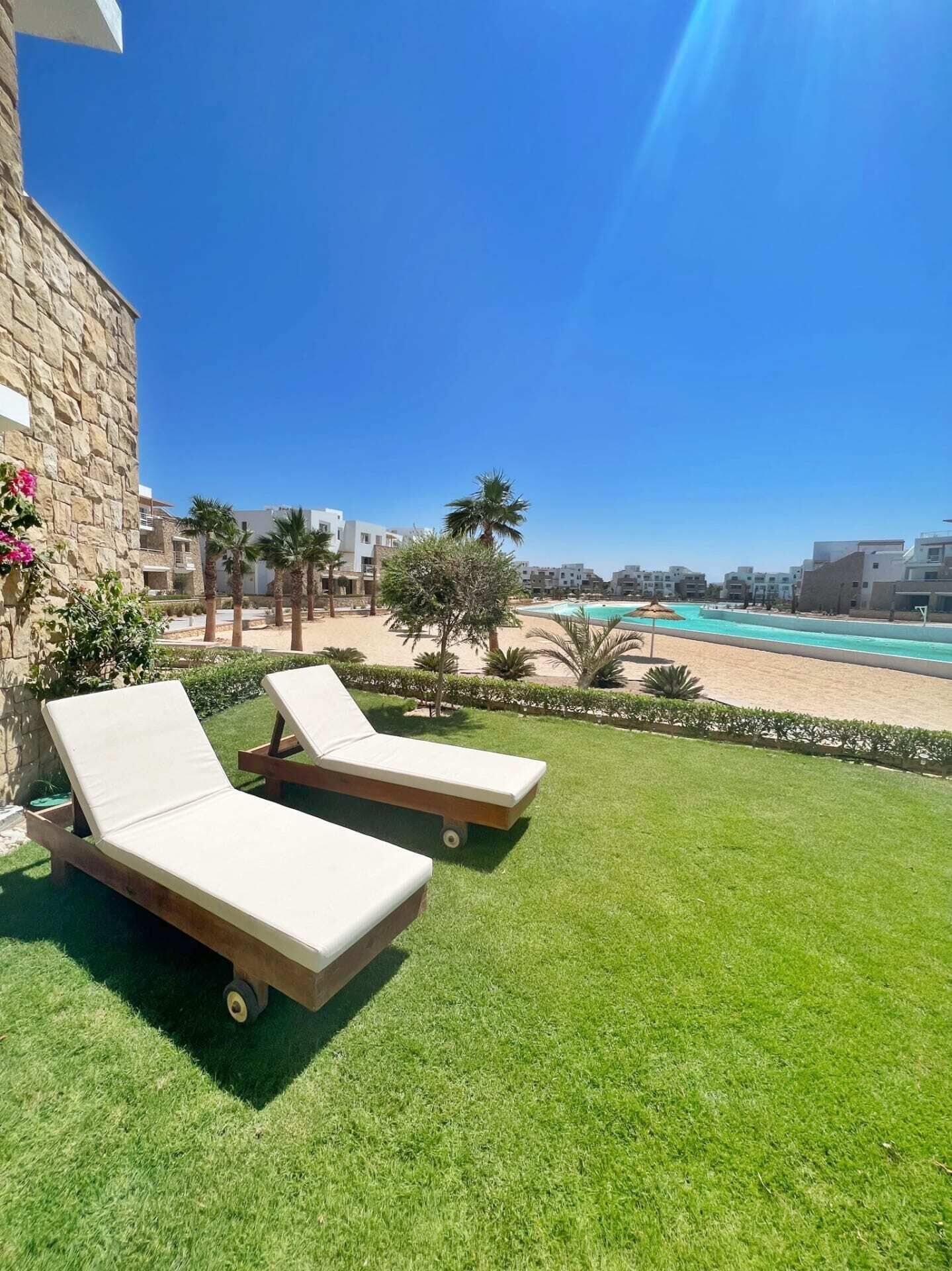 A sunny outdoor area with two lounge chairs, a pool, and a clear view of the beach and buildings in the background.