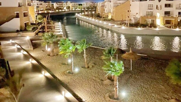 Un hotel frente al agua con piscina, palmeras y un paseo marítimo bien iluminado.