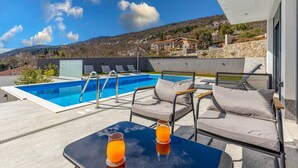 Un espace piscine doté de chaises longues et d’une table, offrant une vue sur une colline parsemée de maisons.