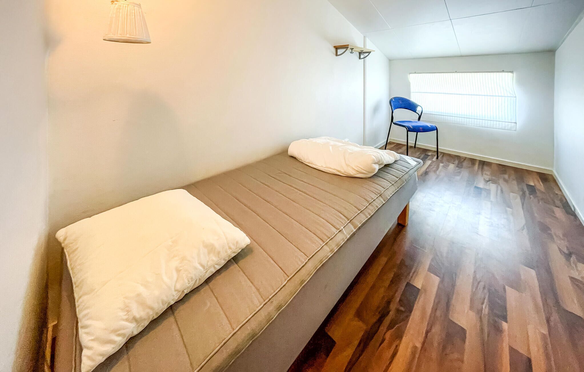 Une petite chambre avec un lit simple, un plancher en bois, un matelas blanc et une chaise bleue.