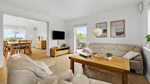 A modern living room with a sofa, a wooden coffee table, and a TV mounted on the wall. There is a dining area with a table and chairs visible through an open door.