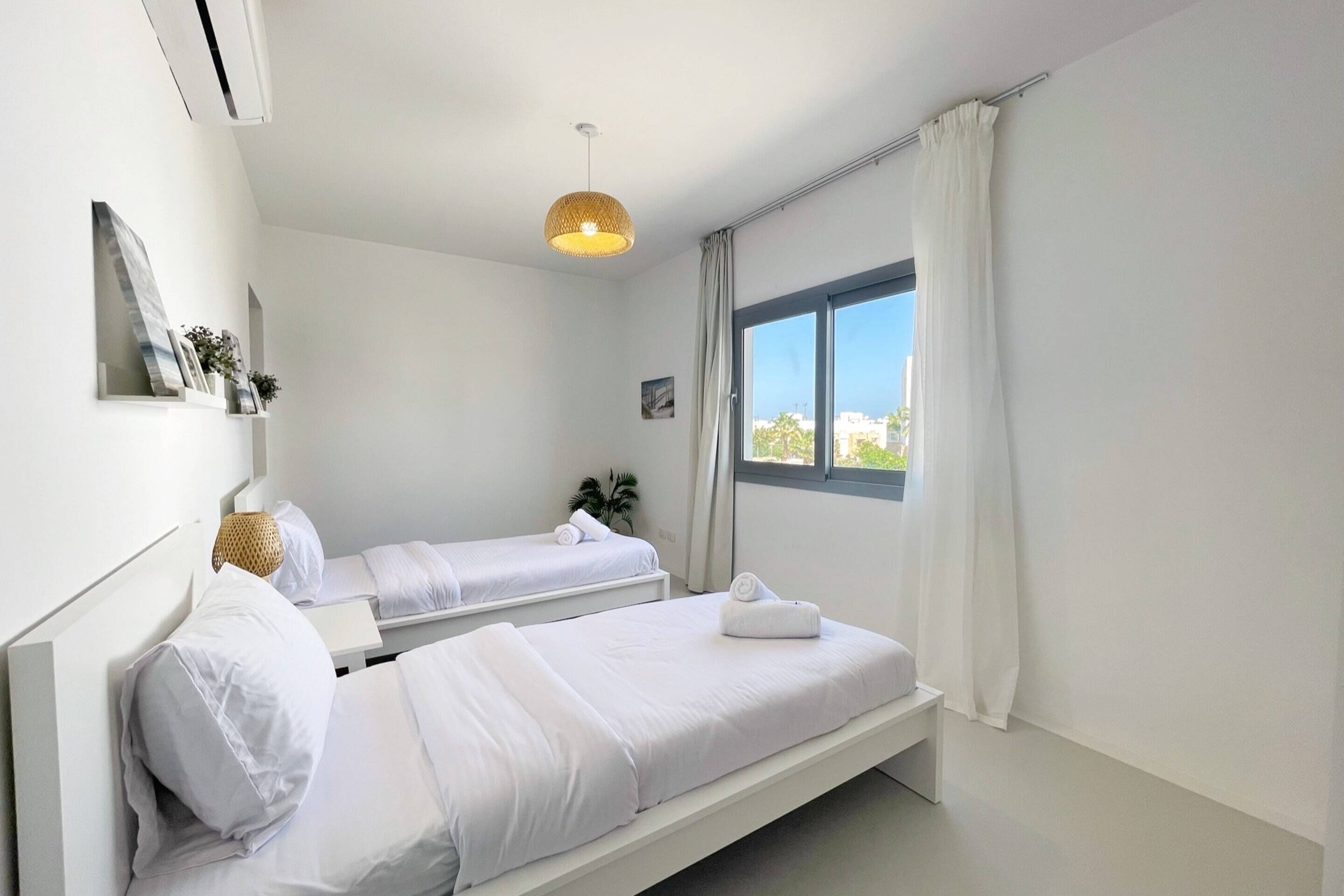 A modern hotel room with two beds, a large window with curtains, a hanging light fixture, and a small plant on the windowsill.
