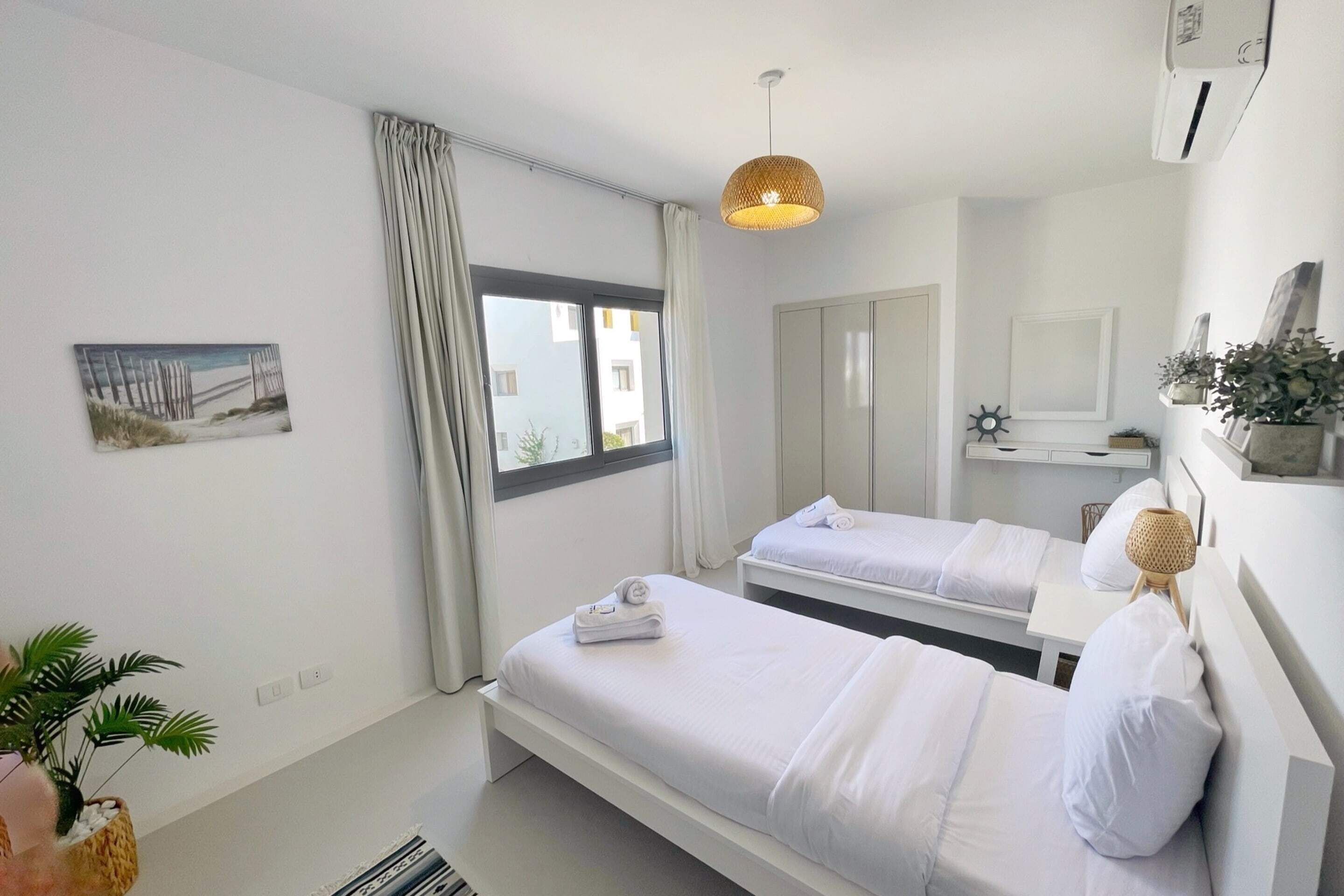 A modern hotel room with two beds, a large window with curtains, a hanging light fixture, and a mirror above the beds.