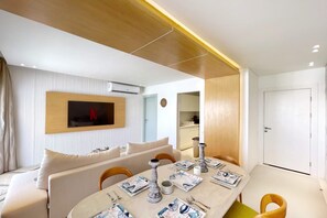 Una sala de estar moderna con un sofá, una mesa de comedor puesta con platos, cubiertos y velas. Se ve un televisor de pantalla plana montado en la pared, y una zona de cocina al fondo.