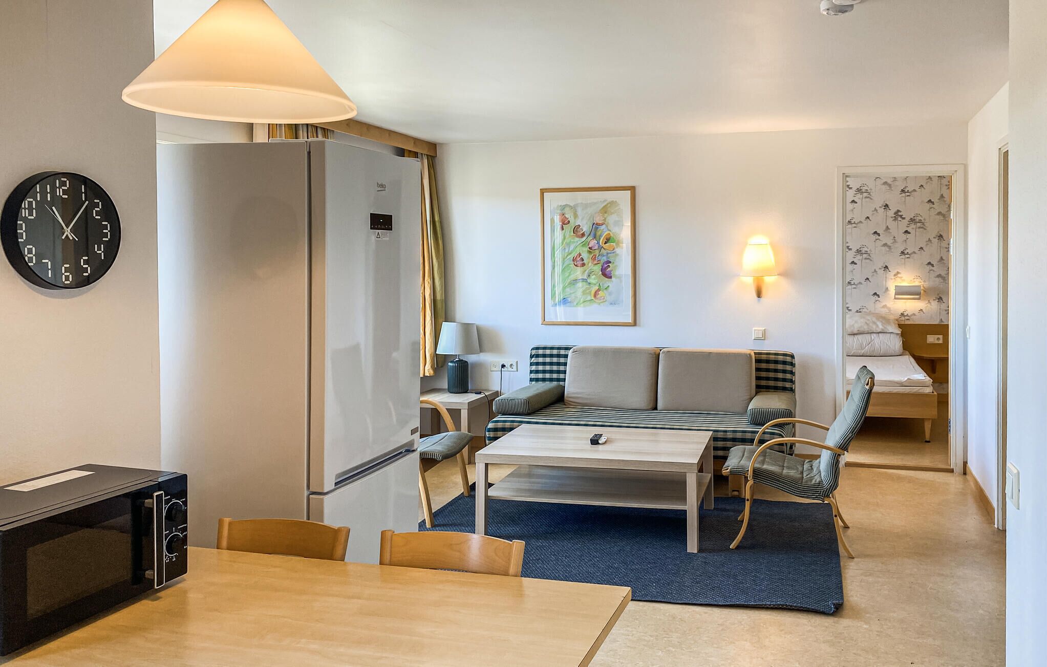 A compact hotel room with a bed, a desk, a chair, a clock, and a microwave.