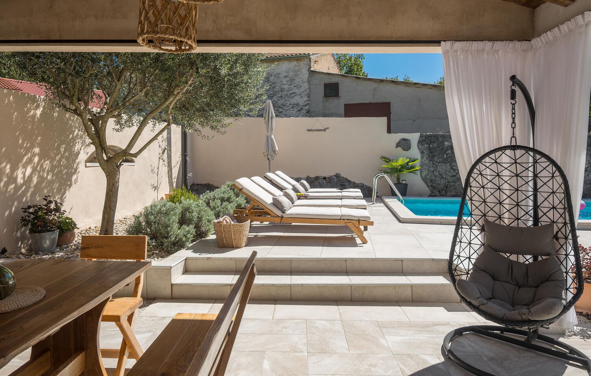 A sunlit outdoor area with a pool, lounge chairs, a wooden table, and a hanging chair.
