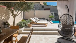 Un espace extérieur ensoleillé avec une piscine, des chaises longues, une table en bois et un fauteuil suspendu.