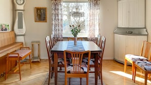 A dining room with a wooden table, chairs, and a bench. There is a chandelier, curtains, and a clock on the wall.