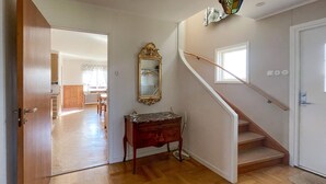 A hallway with a wooden floor, a mirror on the wall, a staircase, and a door leading to another room.