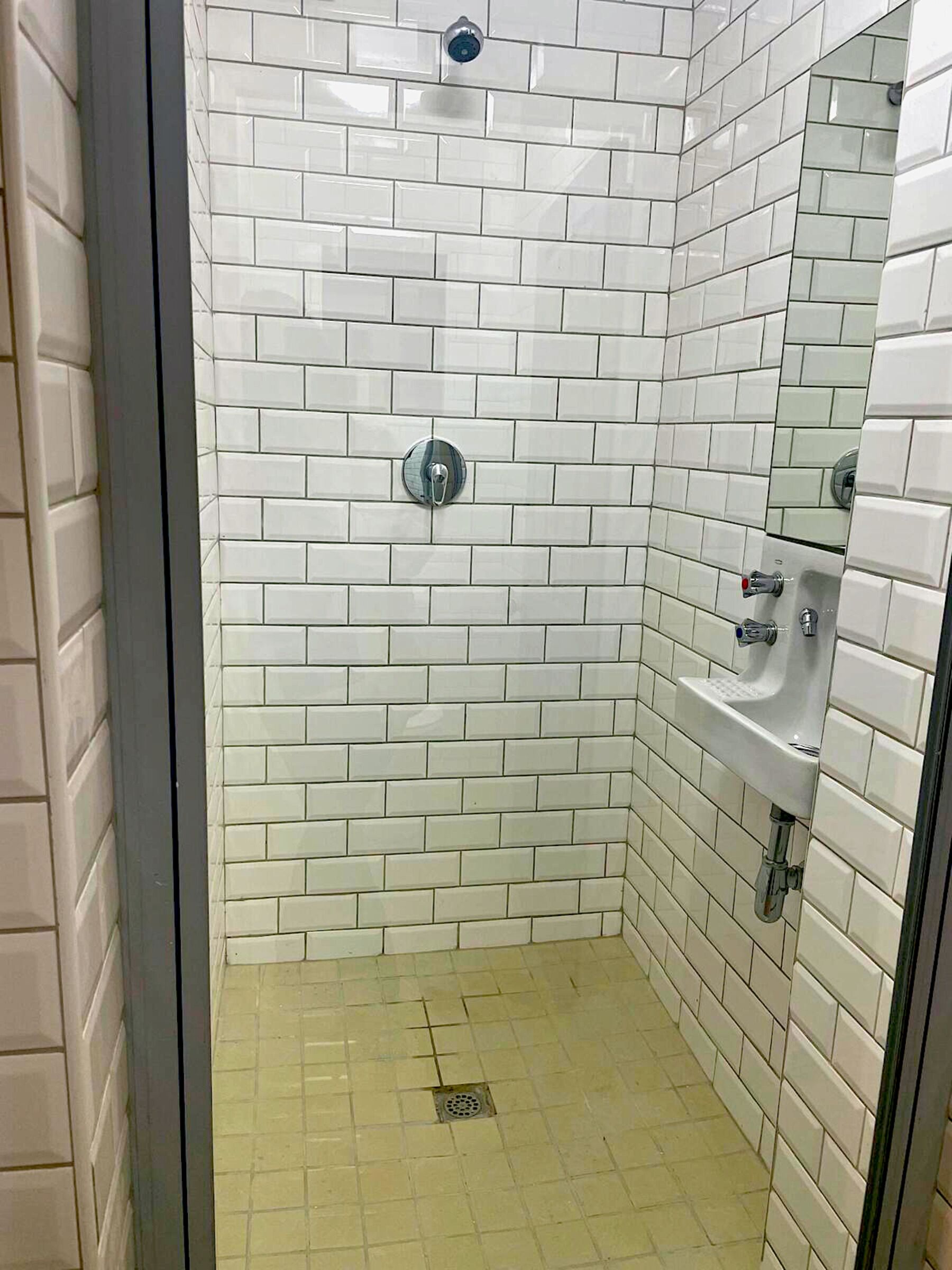 Un petit espace douche fermé, avec des murs et un sol recouverts de carreaux blancs, une porte en verre et un lavabo blanc avec un robinet.