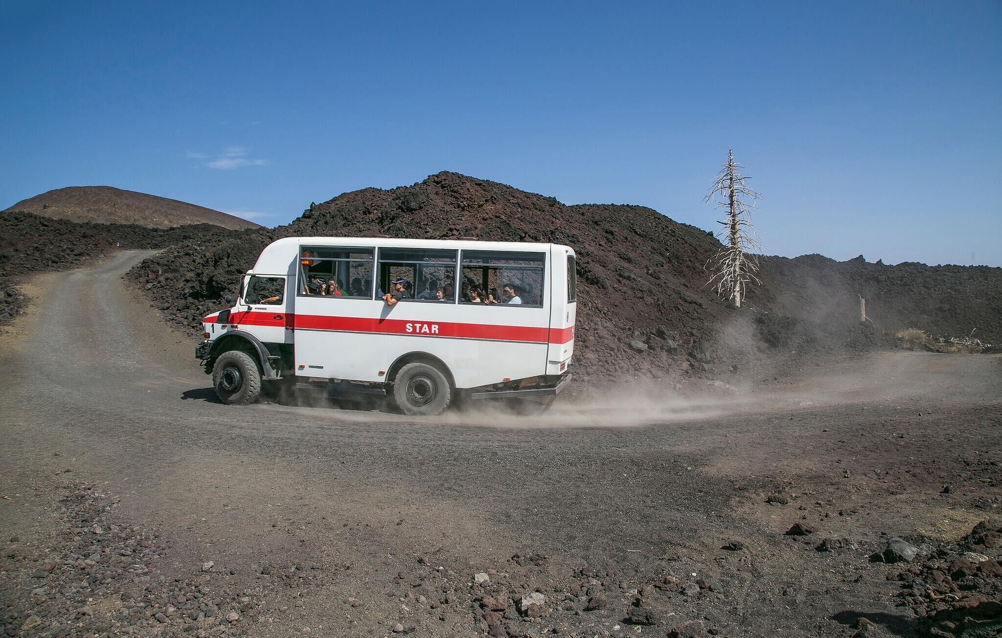 Un autobús blanco con franjas rojas identificado con la etiqueta "STAR" circulando por un camino de tierra, levantando una nube de polvo.