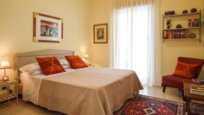 A hotel room with a bed, a nightstand, a chair, a bookshelf, and a window with curtains.