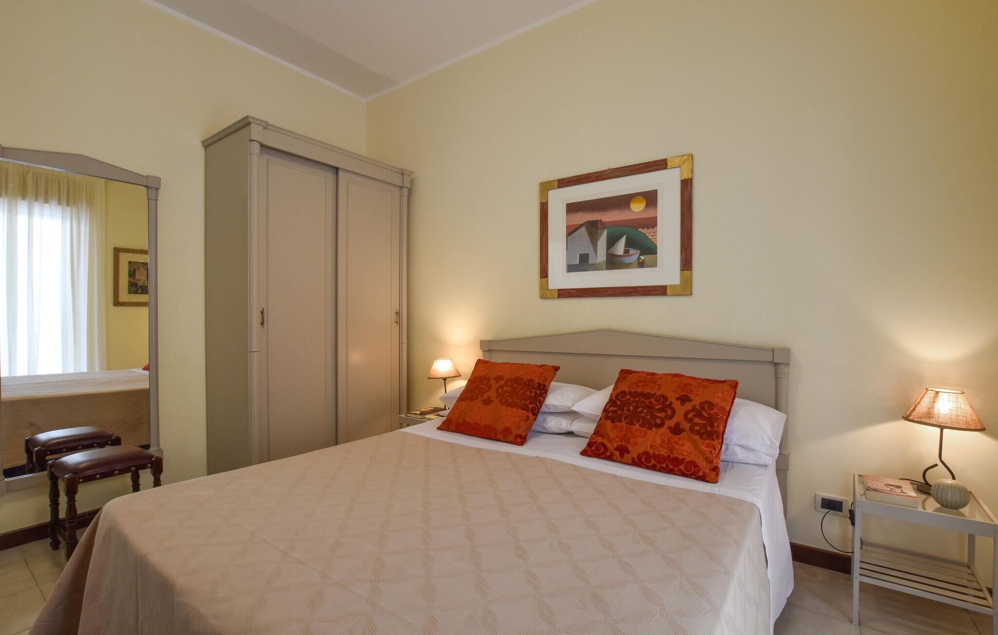 A hotel room with a bed, two pillows, a nightstand with a lamp, a wardrobe, a mirror, and a framed picture on the wall.