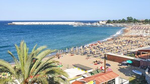 A beach with numerous umbrellas and sun loungers, a clear blue sea, and a distant pier.