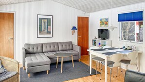Una sala moderna con un sofá seccional gris, una mesita redonda, una mesa de comedor blanca con cuatro sillas, un televisor y una estantería.