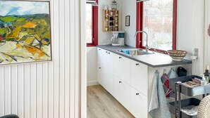 Cocina compacta con armarios blancos, ventana con marco rojo, cafetera y un bol en la encimera.
