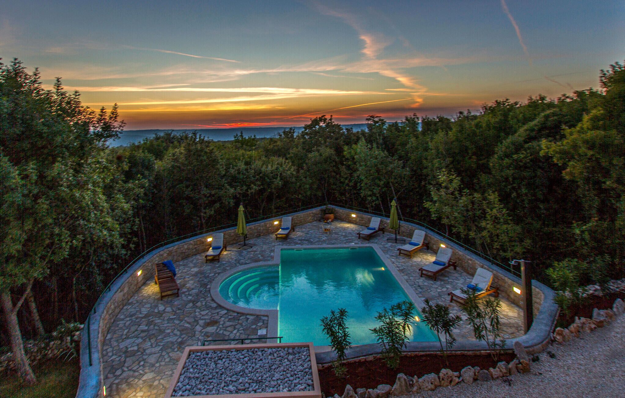 A pool area with lounge chairs and a stone deck, surrounded by trees and overlooking a body of water.