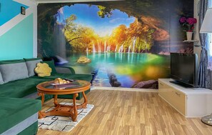 Un salon avec un canapé vert, une table basse, un téléviseur à écran plat et une fresque murale représentant une forêt avec une cascade.
