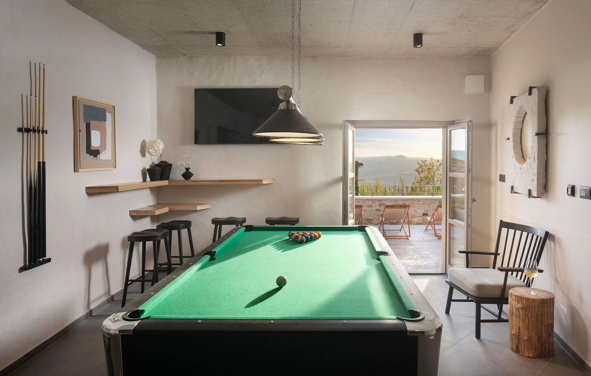 A pool table in a room with a view of the outdoors, a mounted TV, and a wooden shelf.