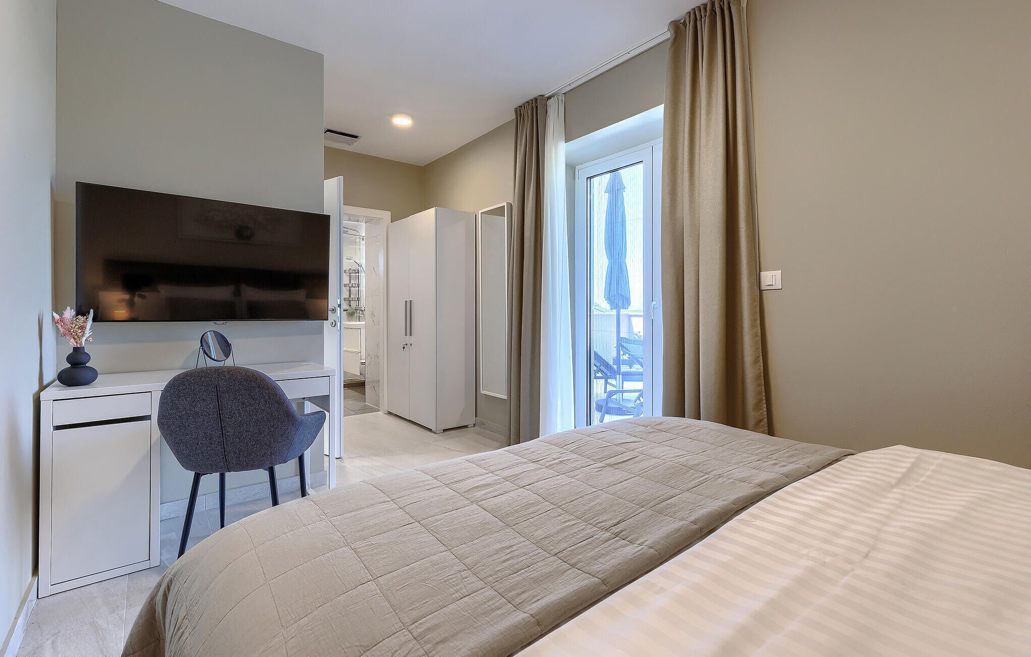 A hotel room with a bed, a desk with a chair, a TV, and a bathroom visible through an open door.