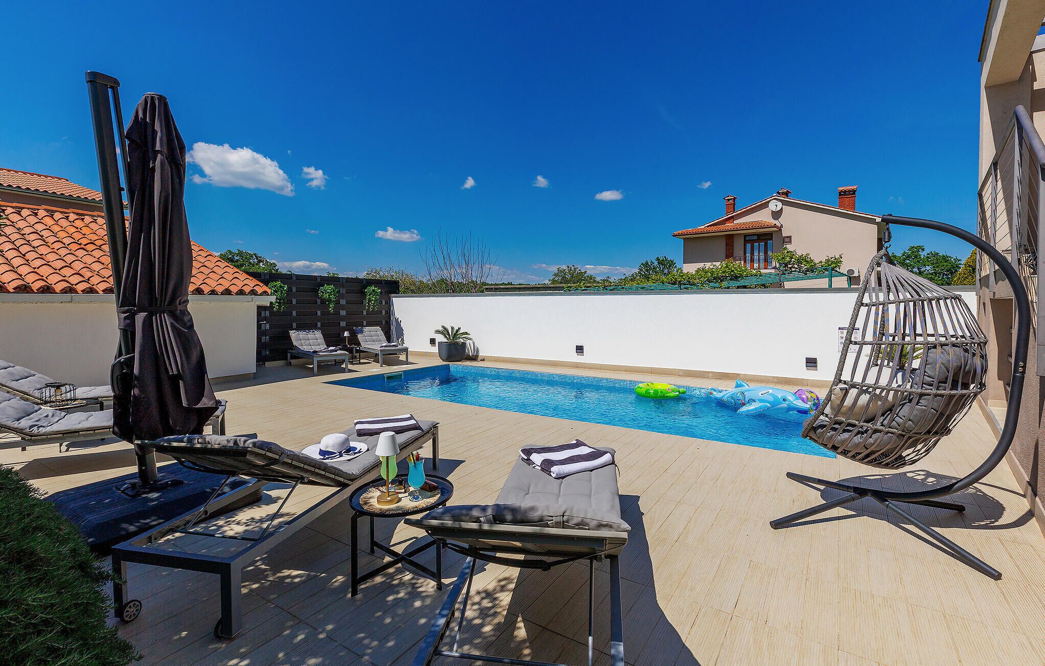 A pool area with lounge chairs, a hanging chair, and a small table with a drink.