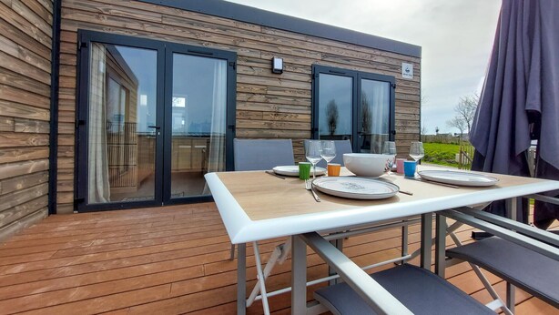An outdoor dining area with a table set for two, a wooden deck, and a modern building with large windows and a balcony. - 2 bedroom beautiful home in Ambon (Ambon)