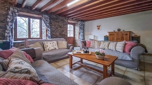 Ein gemütliches Wohnzimmer mit Steinwänden, Holzbalken und einem Steinkamin. Es gibt zwei Sofas, einen Couchtisch und eine Vitrine mit Dekorationen.