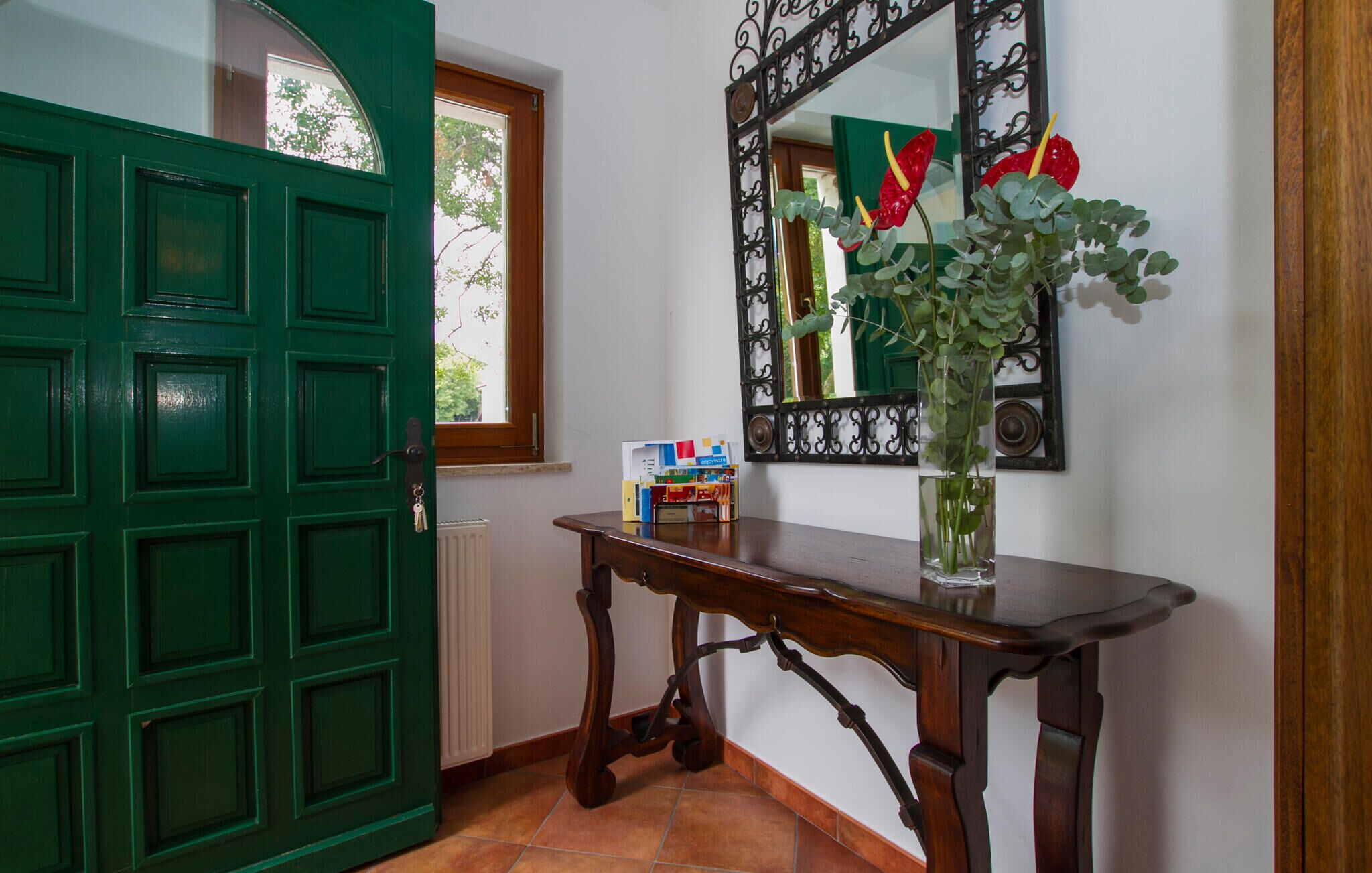 Une porte verte, un miroir avec un cadre décoratif, une table en bois ornée d’un vase contenant des plantes vertes, et une fenêtre donnant sur des arbres.