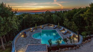 A pool area with lounge chairs and a stone deck, surrounded by trees and overlooking a body of water.