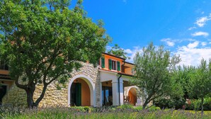 A stone building with a red roof, green shutters, and a garden with lavender.