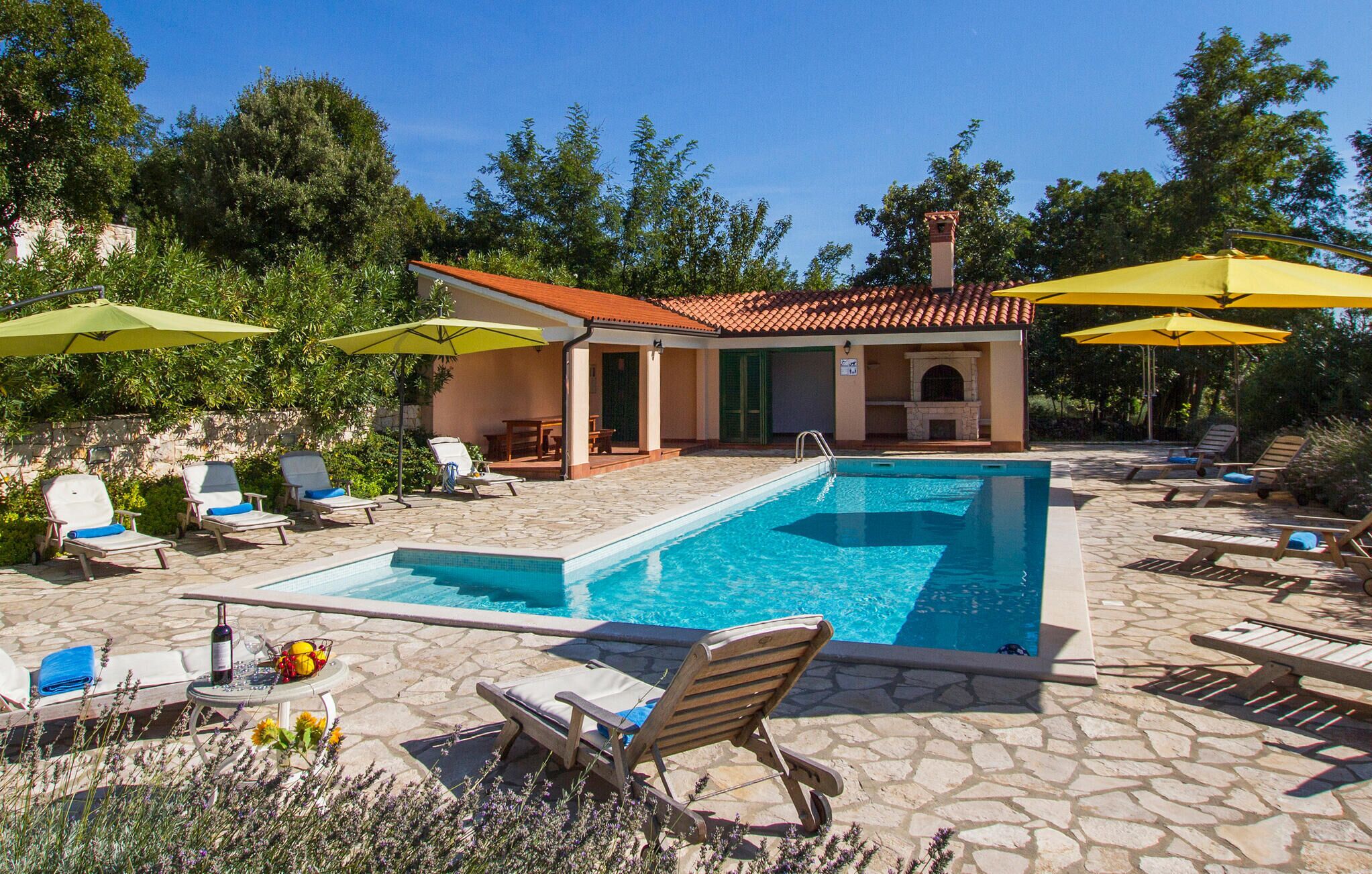 Un espace piscine avec des chaises longues et des parasols, une terrasse en pierre et un bâtiment doté d’un toit en tuiles.