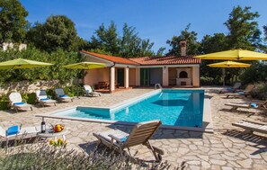 Un área de piscina con sillones de descanso y sombrillas, un patio de piedra y un edificio con tejado de baldosas.