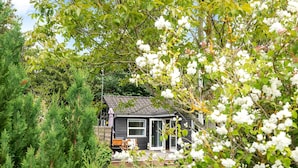 Une maison traditionnelle de style japonais, avec un toit de chaume, entourée d’une végétation luxuriante et d’arbres en fleurs.