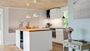 Une cuisine moderne avec des meubles blancs, un îlot central en bois, des luminaires suspendus et une vue sur l’extérieur.