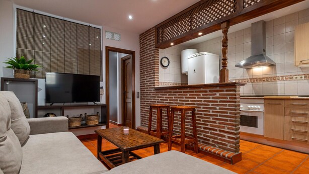 A living room with a TV, sofa, and a small table. There is a kitchen area with a stove and cabinets.