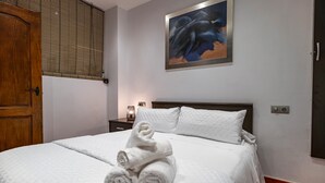 A hotel room with a bed, a painting, a wooden door, and a nightstand with a lamp.