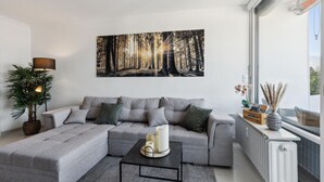A modern living room with a grey sectional sofa, a coffee table with candles, a floor lamp, and a large forest-themed artwork on the wall.