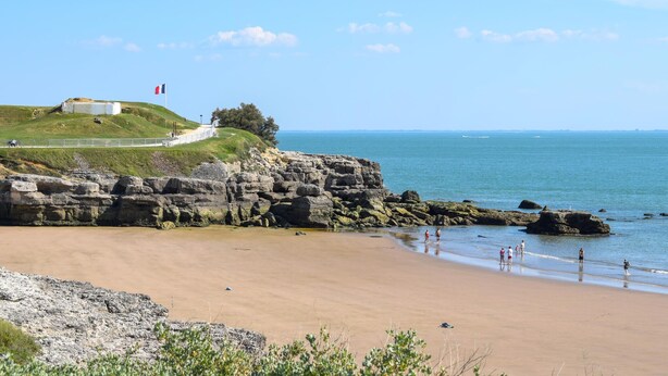 Eine Küstenlandschaft mit Sandstrand, felsigen Klippen und einem grasbewachsenen Hügel mit einer Flagge.